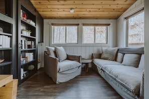 Ein gemütliches Zimmer mit Holzbalkendecke, zwei Fenstern mit Jalousien, einem Bücherregal, einem Sofa und einem Sessel.