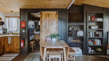 A wooden interior with a kitchen area, a dining table with chairs, and a bookshelf.