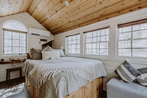 Ein gemütliches Schlafzimmer mit Holzbalkendecke, einem großen Bett mit weißer Bettdecke, mehreren Fenstern und einer Sitzbank am Fenster.
