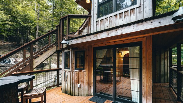 A wooden cabin with a deck, a staircase, and a large sliding glass door. - The Beech Treehouse | Hocking Hills (South Bloomingville)