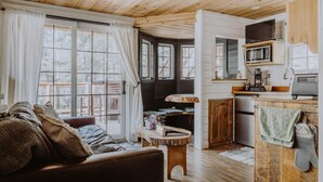 L’intérieur chaleureux d’un chalet avec un plafond en bois, un espace de vie comprenant un canapé et une table basse, une cuisine équipée d’un four à micro-ondes et d’un évier, et de grandes fenêtres avec des rideaux.