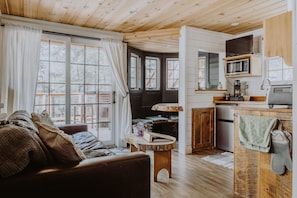 Ein gemütliches Blockhaus-Interieur mit Holzbalkendecke, einem Wohnbereich mit Sofa und Couchtisch, einer Küche mit Mikrowelle und Spüle sowie großen Fenstern mit Vorhängen.