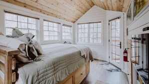A cozy bedroom with a wooden ceiling, large windows, a bed with a quilted blanket, and a wooden nightstand. - The Beech Treehouse | Hocking Hills (South Bloomingville)