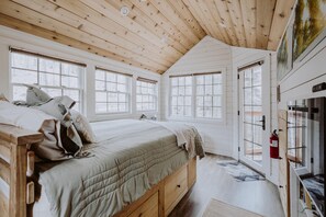 A cozy bedroom with a wooden ceiling, large windows, a bed with a quilted blanket, and a wooden nightstand. - The Beech Treehouse | Hocking Hills (South Bloomingville)
