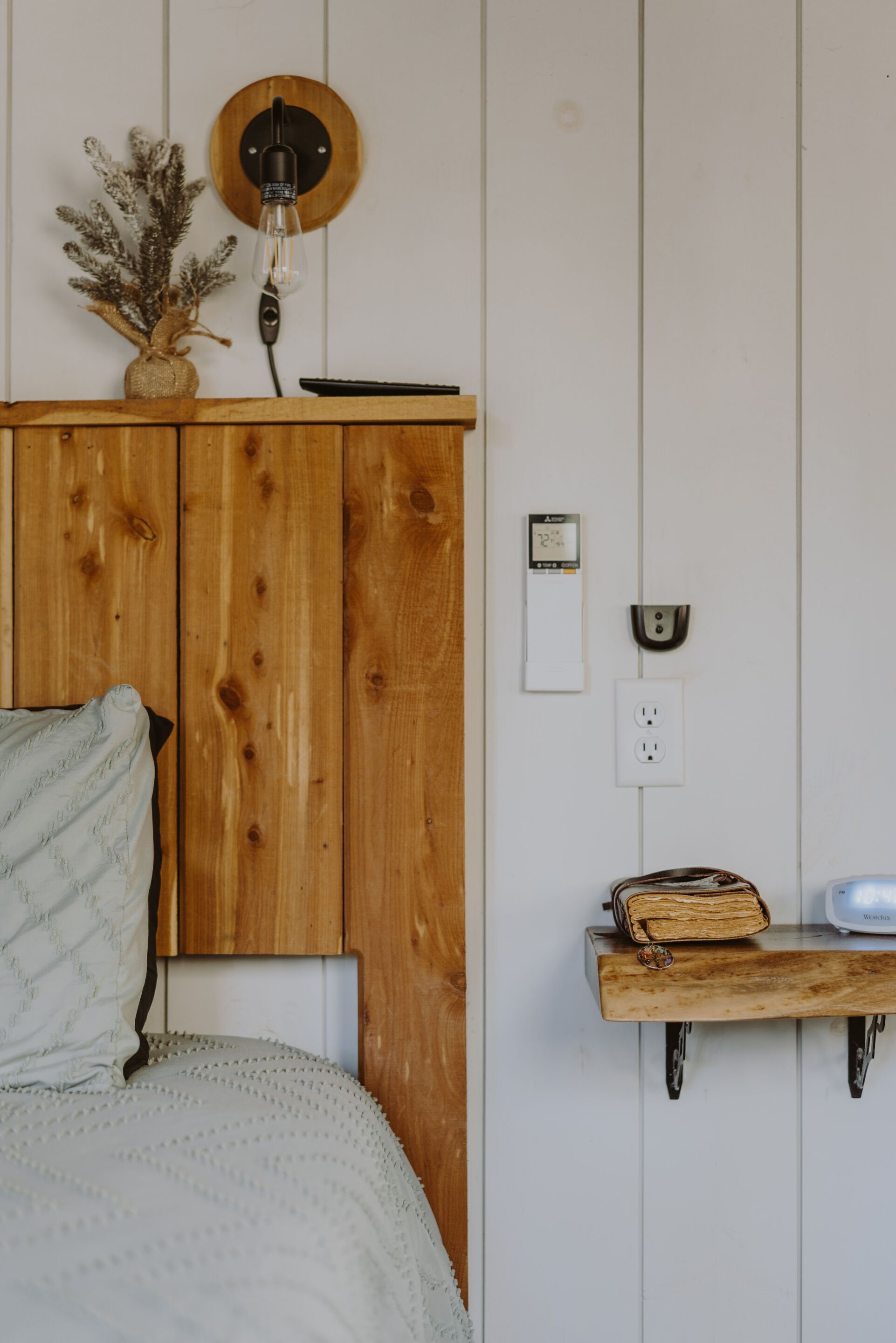 A bedroom with a wooden headboard, a bedside lamp, a small shelf with a book and a toaster, and a wall clock.