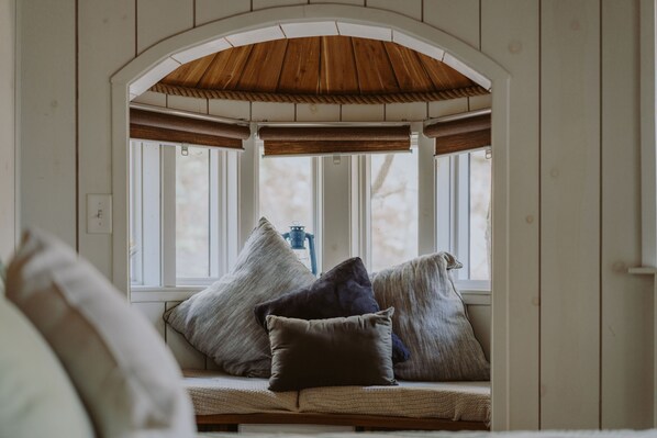 Un coin salon accueillant avec un banc et des coussins, un plafond en bois et une vue sur l’extérieur à travers une fenêtre munie de stores.