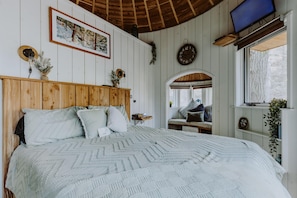 Ein Schlafzimmer mit einem hölzernen Kopfteil, einem Bett mit einem gemusterten Überwurf, einer Wanduhr, einem Fernseher und einem Fenster mit Blick ins Freie.