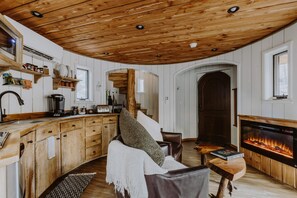 A cozy cabin interior with wooden ceilings, a fireplace, a dining area, and a kitchen with wooden cabinets and shelves.