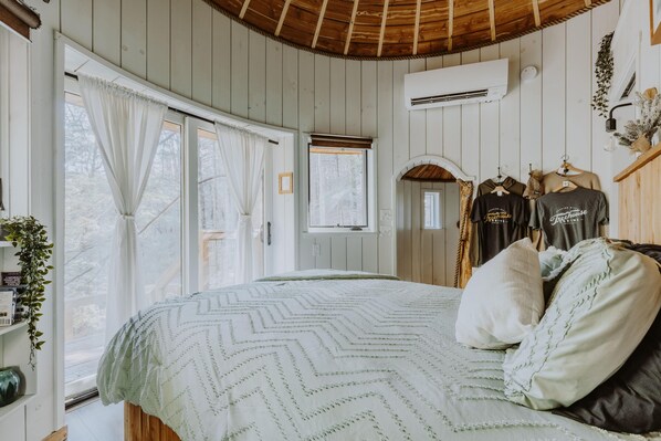 A bedroom with a bed, a window, an air conditioning unit, and a clothing rack with t-shirts.
