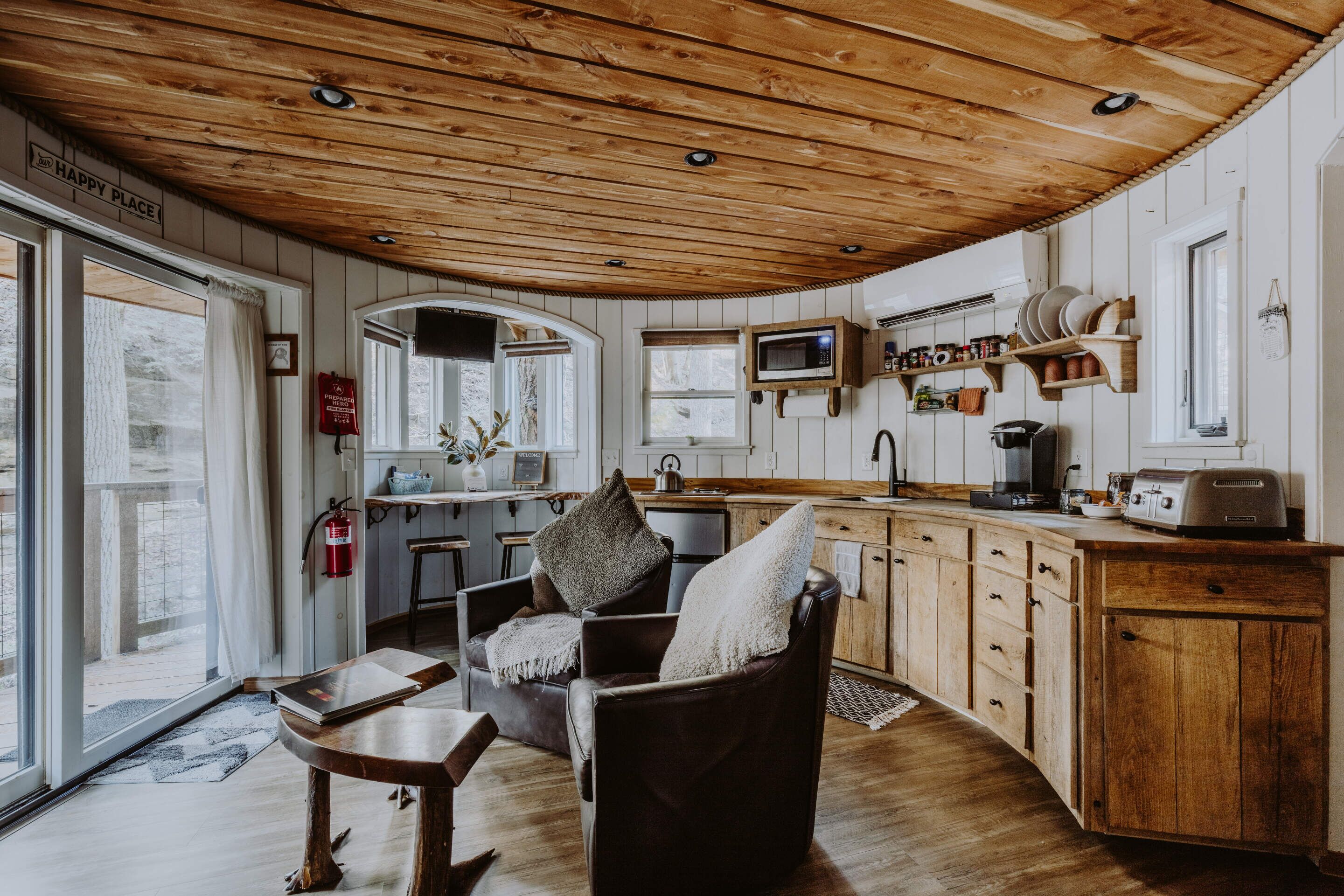 A cozy kitchen with wooden cabinets, a microwave, and a toaster. There is a chair with a cushion and a table with a book.