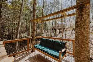 Une terrasse en bois avec un fauteuil suspendu et une vue sur une cabane dans les arbres, située au cœur d’une forêt.