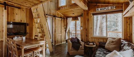 A wooden cabin interior with a dining area, a sofa, and a window with a view of trees.