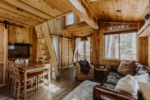 A wooden cabin interior with a dining area, a sofa, and a window with a view of trees.