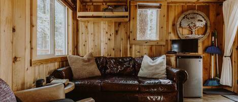A cozy cabin interior with a leather couch, a small refrigerator, and a window with a view of trees.