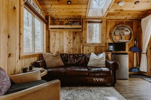 A cozy cabin interior with a leather couch, a small refrigerator, and a window with a view of trees.