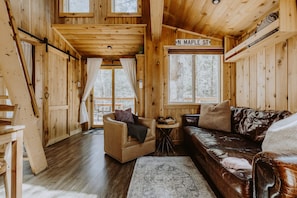 Interior acogedor de una cabaña con detalles de madera, un sofá de cuero, una pequeña mesa redonda y un letrero que dice 'N. Maple St'.