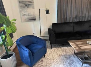 Una sala de estar con un sillón azul, un sofá negro, una mesa de centro de madera y una lámpara de pie.