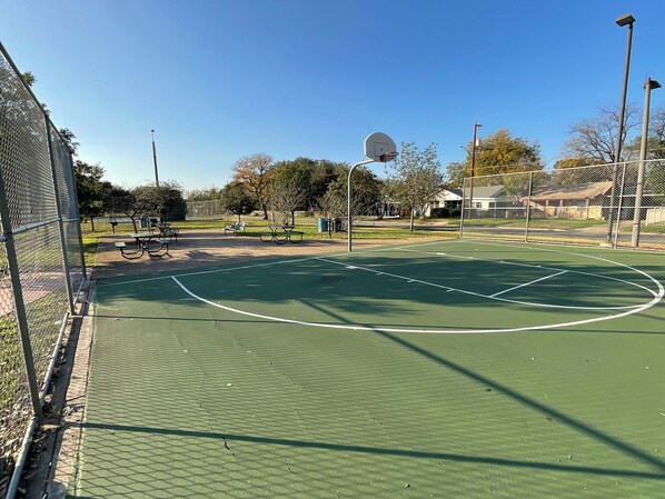 Una cancha de baloncesto al aire libre con una valla, bancos y árboles de fondo.