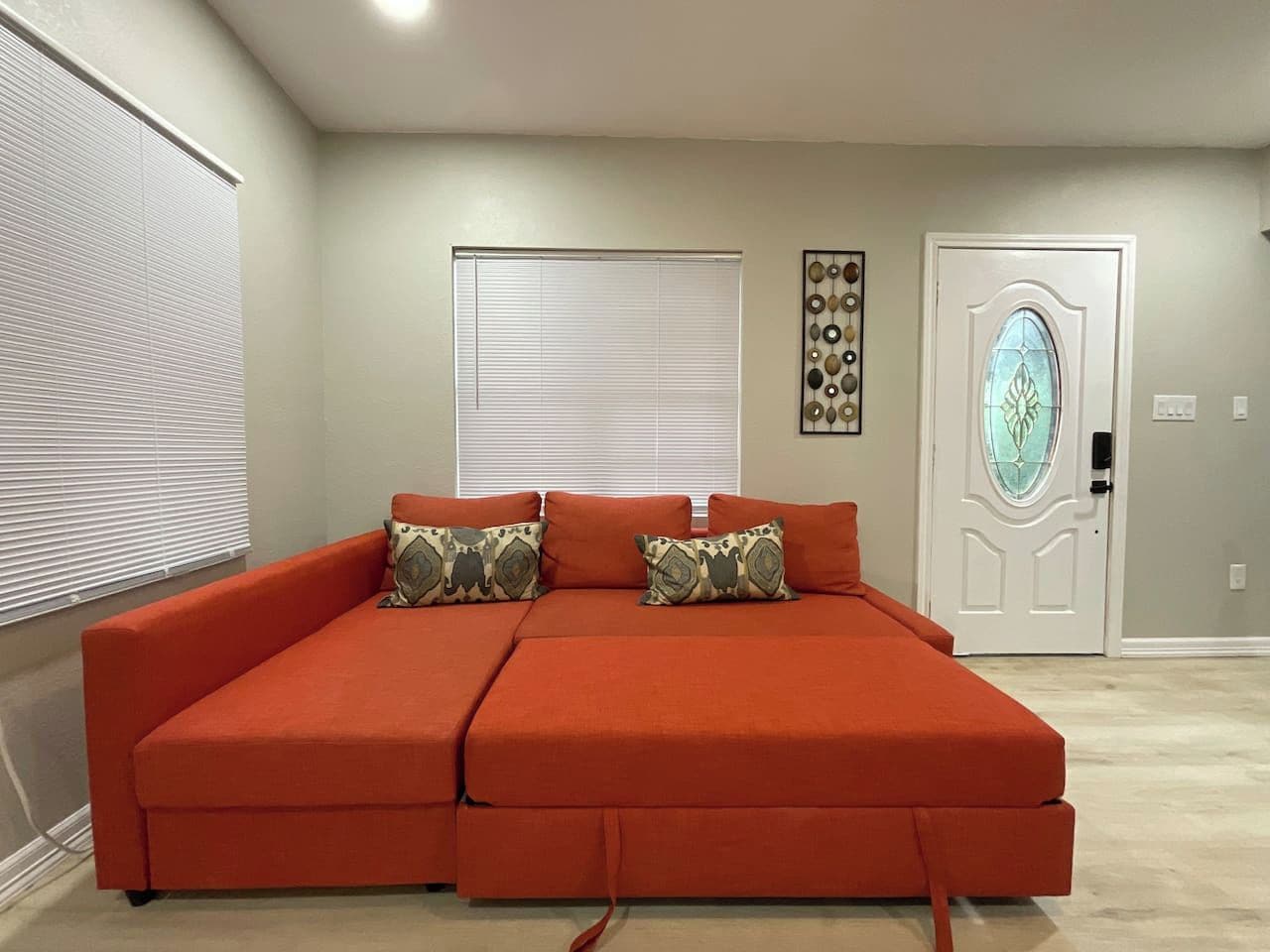 Habitación con un sofá grande color naranja, dos ventanas con persianas, un adorno de pared y una puerta con panel de vidrio.