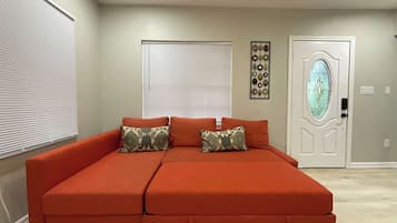 Habitación con un sofá grande color naranja, dos ventanas con persianas, un adorno de pared y una puerta con panel de vidrio.