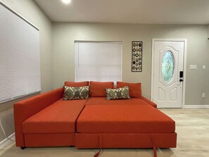 A room with a large orange sofa, two windows with blinds, a decorative wall piece, and a door with a glass panel.
