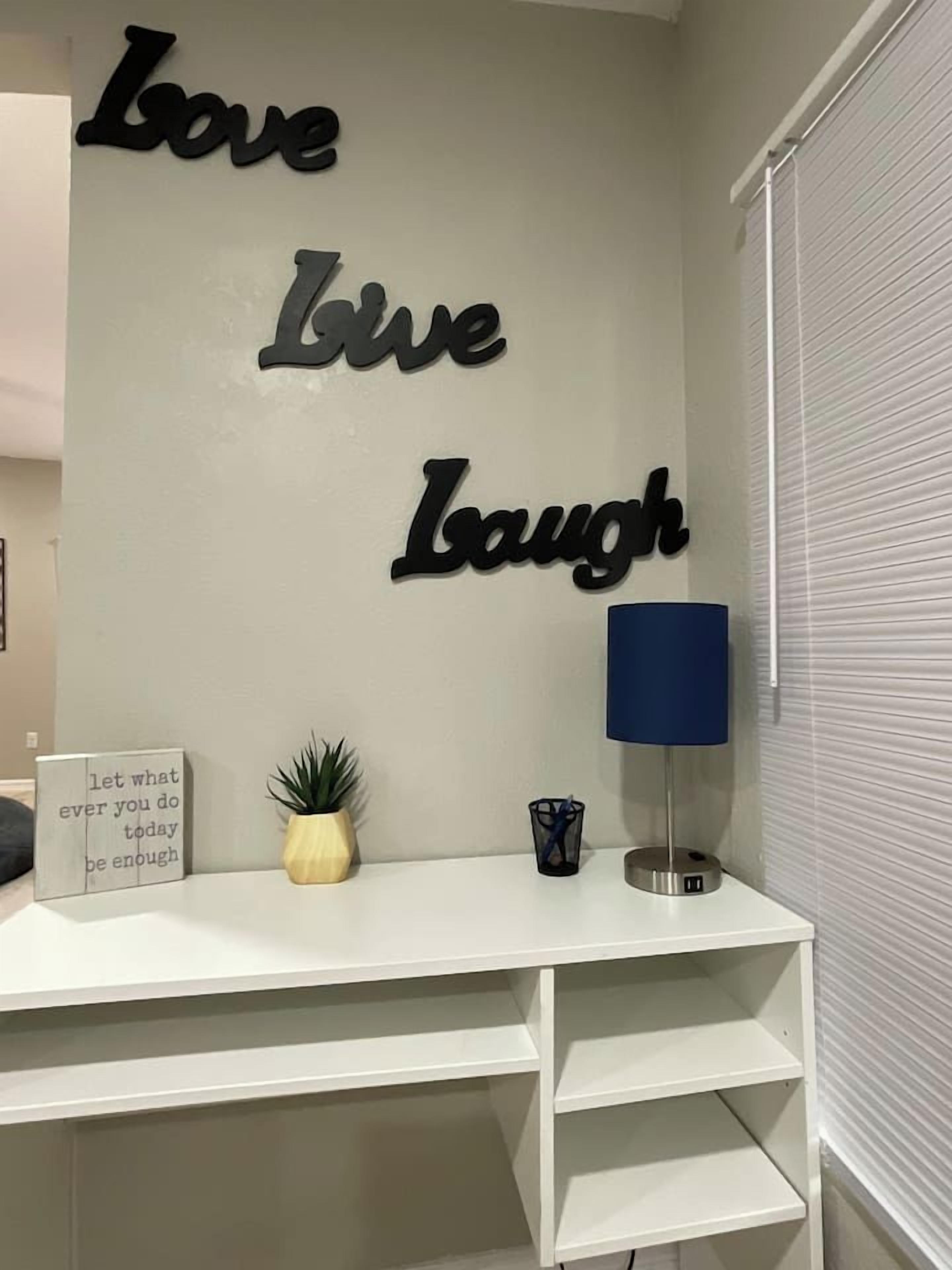 Quarto com escrivaninha branca, parede com frases motivacionais, uma pequena planta, abajur azul e um letreiro com uma citação inspiradora.
