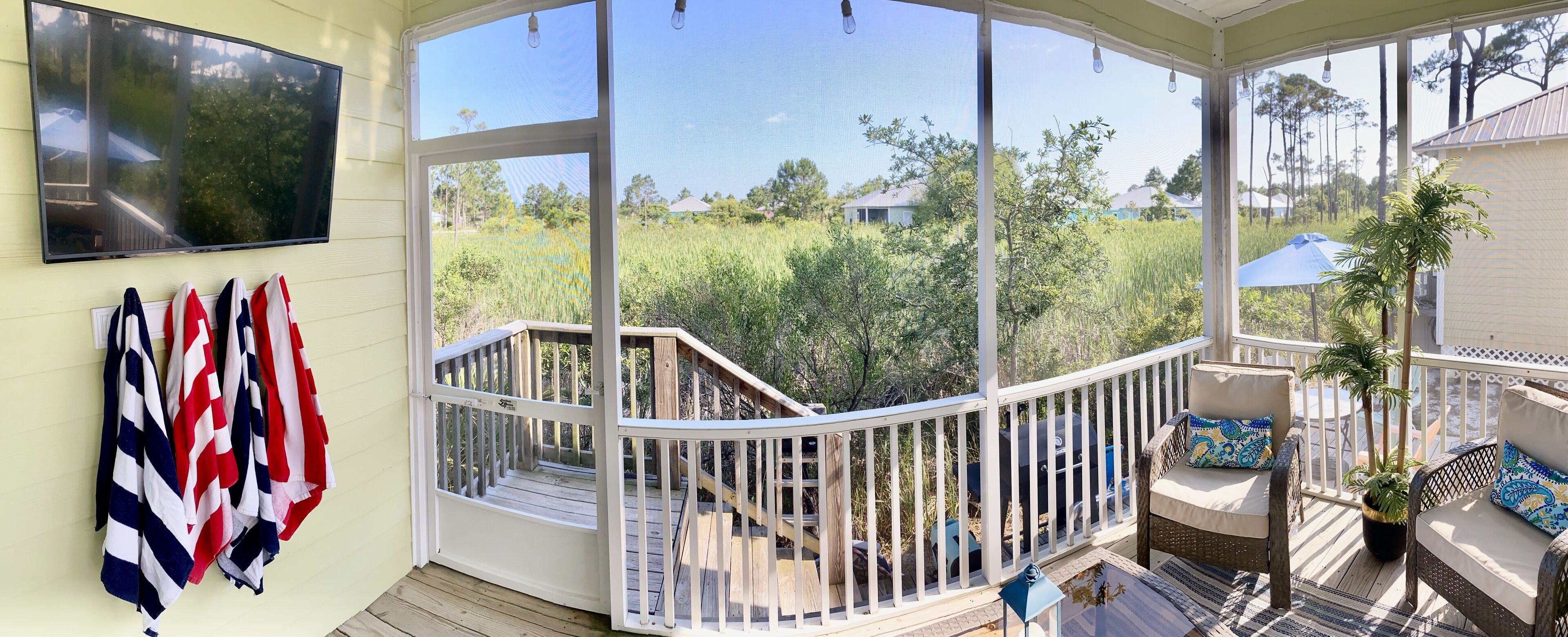 A covered outdoor deck with a wooden railing, a television mounted on the wall, and a view of a grassy area with trees and houses in the background.