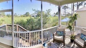 A covered outdoor deck with a wooden railing, a television mounted on the wall, and a view of a grassy area with trees and houses in the background. - Beach Cottage/2 pools/Golf Cart Friendly/Beach Access (Gulf Shores)