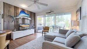 A modern living room with a flat-screen TV, wooden wall paneling, a ceiling fan, and a large window offering a view of the outdoors.