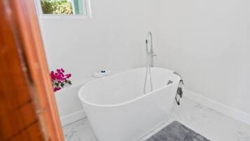 A modern bathroom with a freestanding bathtub, a small window, and a decorative flower arrangement.
