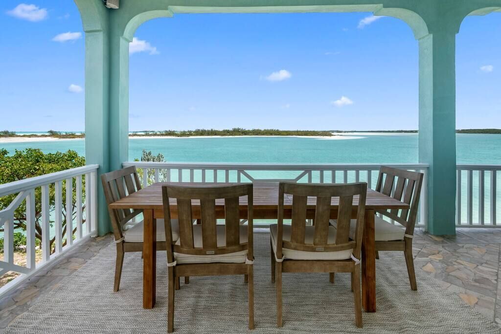 A covered outdoor seating area with a table and four chairs, overlooking a beach with clear turquoise waters and a distant shoreline.