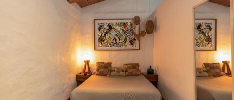 A bedroom with a wooden bed, two bedside tables with lamps, a large mirror, and two framed tropical artwork pieces.