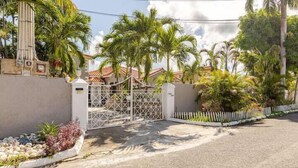 A gated residential area with a paved driveway, palm trees, and a white fence. - The cloud room At Csompo Silver Manor (Tower Isle)