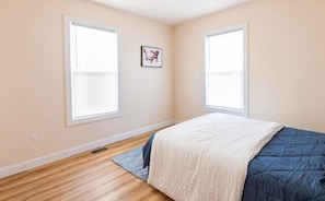 Quarto com cama, duas janelas, parede em tom claro e chão de madeira.