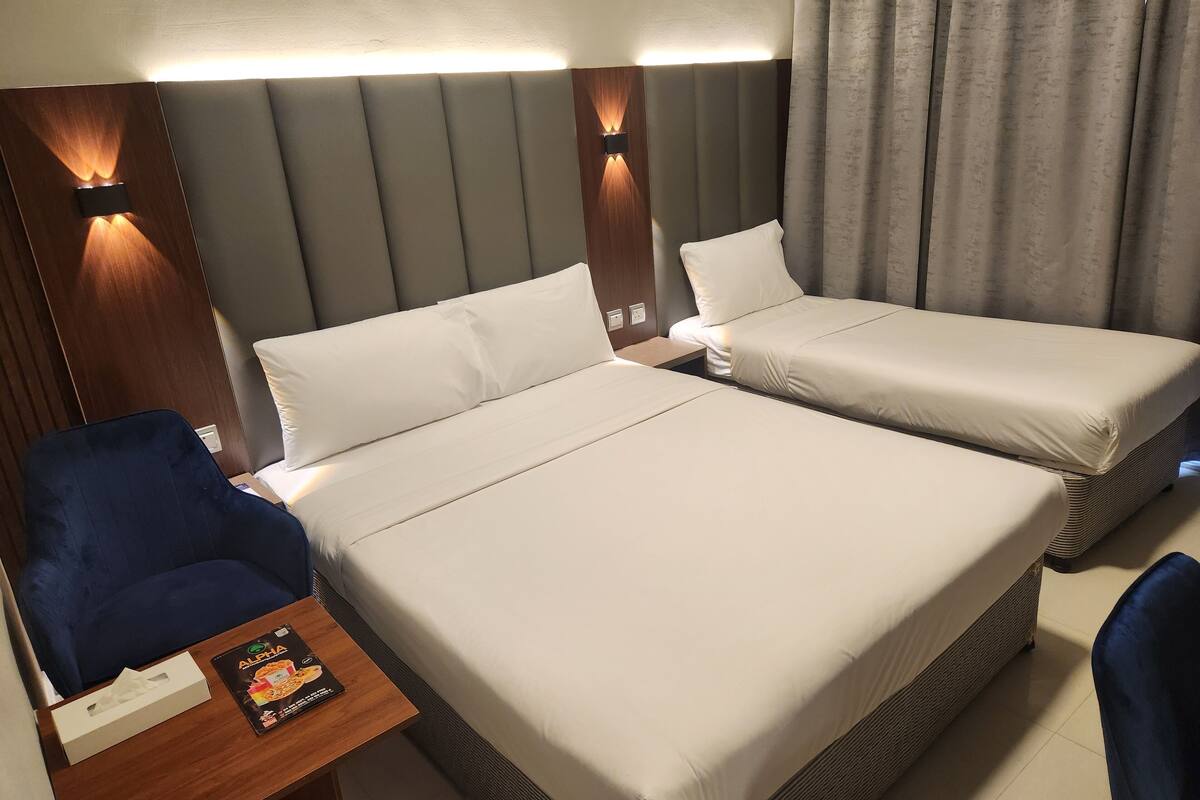A hotel room with two beds, a blue chair, a small table with a tissue box and a magazine, and a curtain.