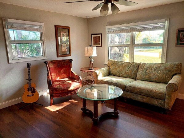Uma sala de estar com piso de madeira, mesa de centro de vidro, poltrona vermelha, sofá verde e uma guitarra.