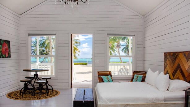 A room with a bed, a small table, and a bench. There are two windows with views of palm trees and the ocean.