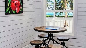 A room with a round table and four chairs, a large window with a view of palm trees and the ocean, and a painting of a red flower on the wall.