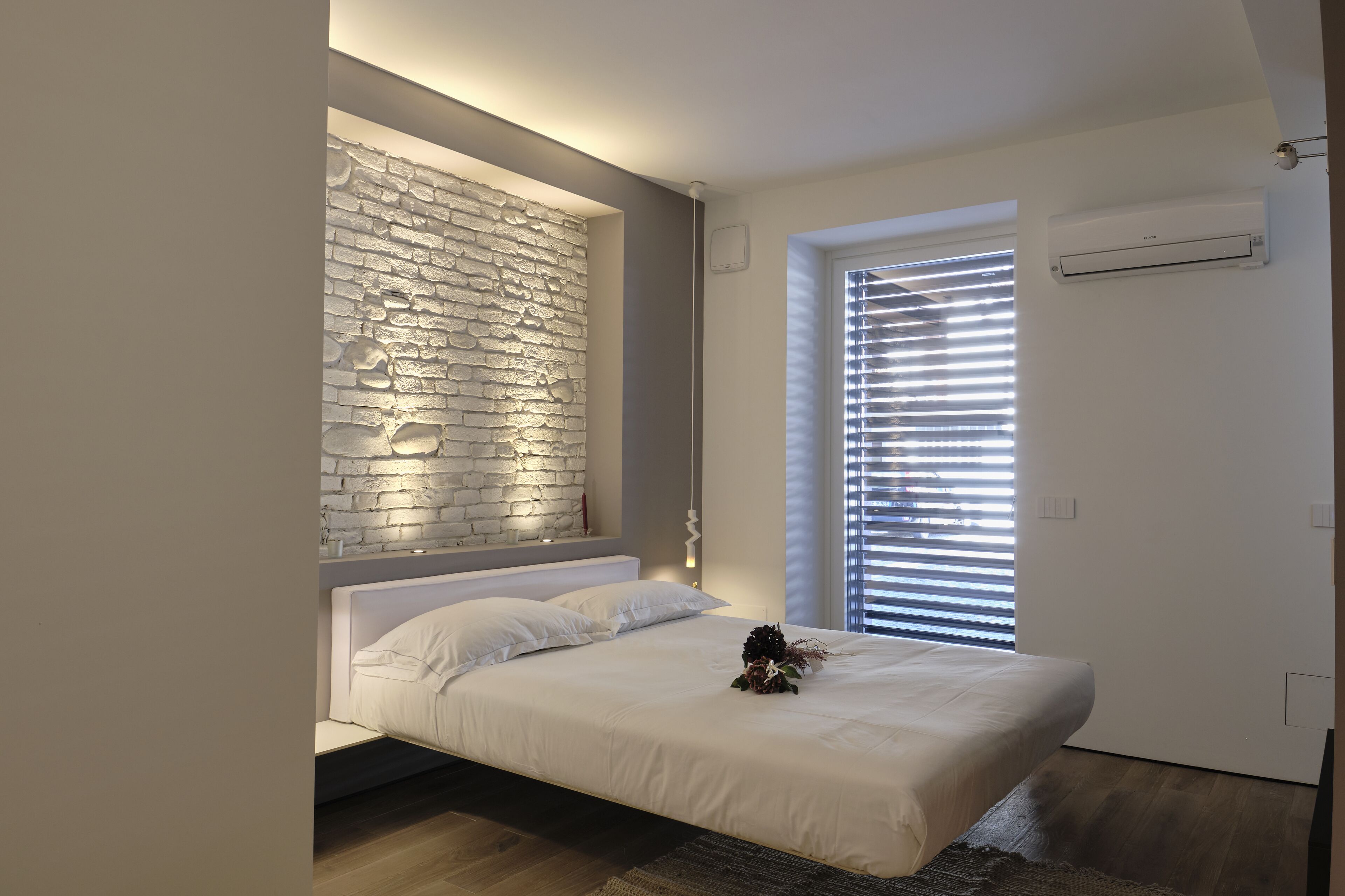 Une chambre moderne avec un lit blanc, un mur en pierre, une fenêtre avec des stores et un appareil de climatisation.