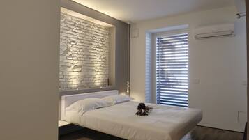 Une chambre moderne avec un lit blanc, un mur en pierre, une fenêtre avec des stores et un appareil de climatisation.