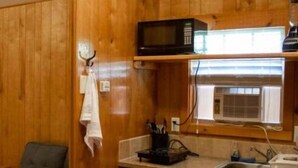 A compact kitchen with wooden walls, a microwave, a sink, and a window with an air conditioning unit.