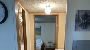 Een smalle gang die leidt naar een slaapkamer met een bed, een nachtkastje en een lamp. Er hangt een klok aan de muur en er staat een stoel in de hoek.