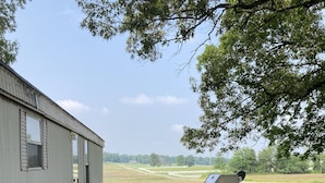 Een houten terras dat uitkomt op een grasveld, met een barbecue en een metalen gebouw op de achtergrond.