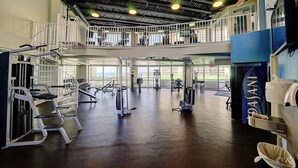 A multi-level gym with various exercise machines, a mezzanine level with a railing, and large windows offering views.