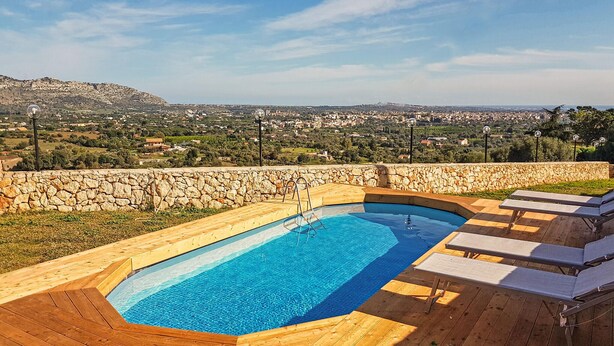 A rectangular swimming pool with a stone wall and wooden deck, surrounded by lounge chairs and a scenic view of a town and hills. - Cozy home in Floridia (Floridia)
