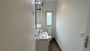 A narrow, white-walled bathroom with a white sink, mirror, and toilet. - Oasis Urbain (Argenteuil)