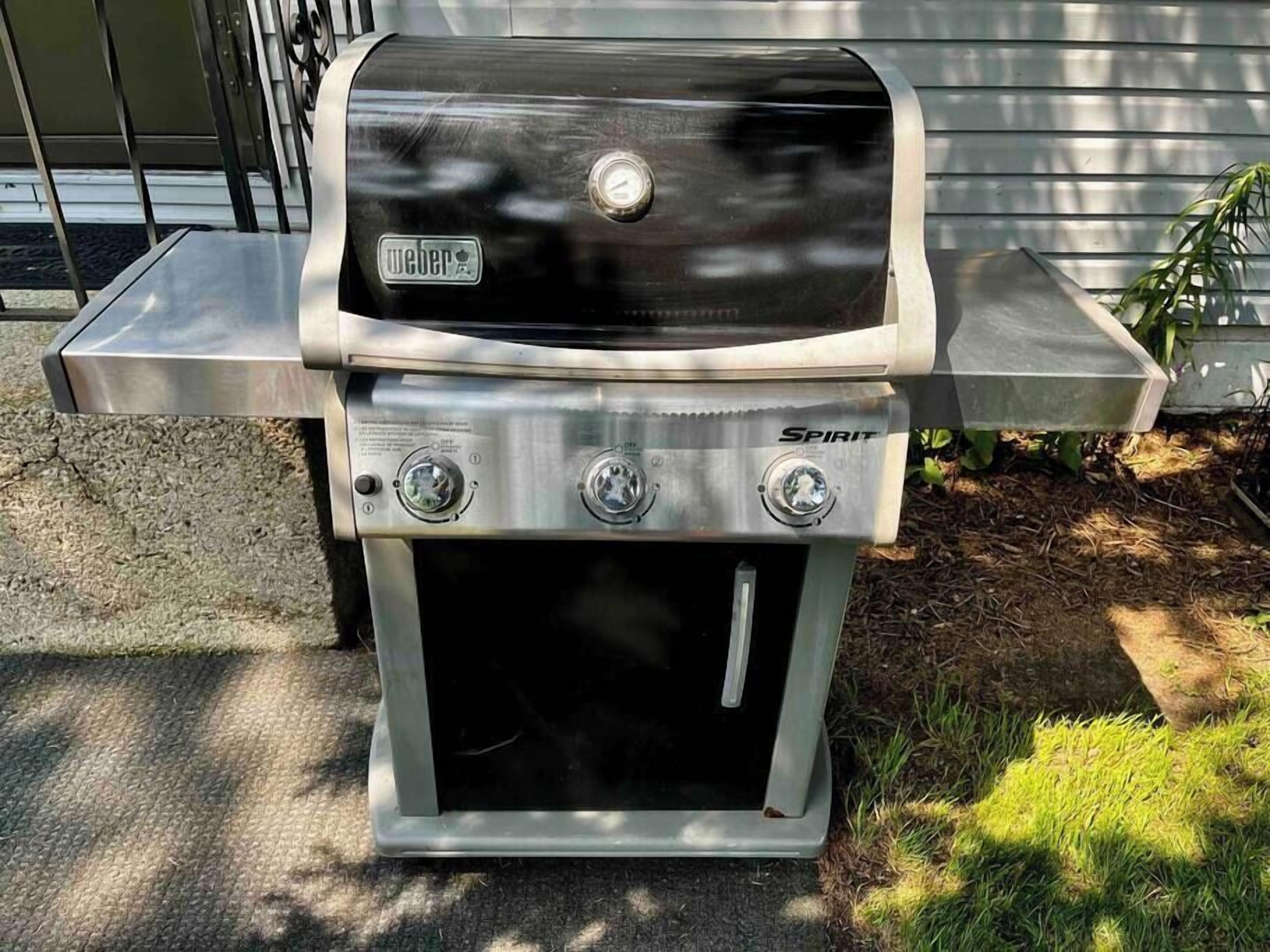 Un barbecue Weber Spirit portable, doté d’une finition en acier inoxydable, placé à l’extérieur, avec une rambarde en métal et de la verdure en arrière-plan.