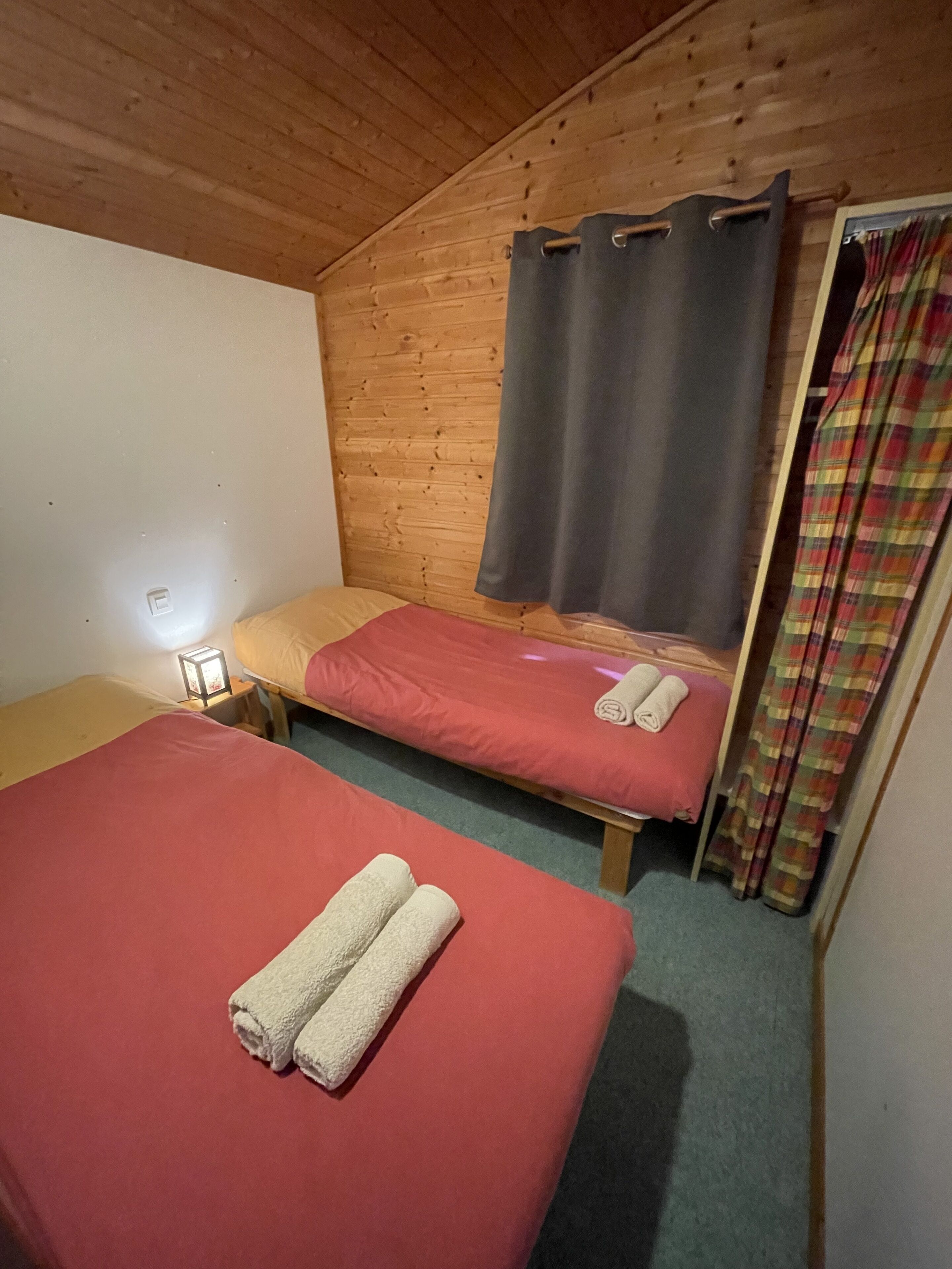 Una habitación pequeña y acogedora con dos camas individuales, paredes de madera, un cortinaje y una cortina escocesa.