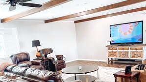 Un salón con un sofá de cuero marrón, una mesa redonda, un televisor sobre un mueble de madera y un ventilador de techo.