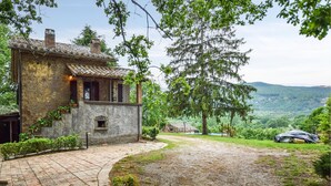 A rustic stone house with a tiled roof, a stone staircase, and a paved driveway leading to a grassy area.